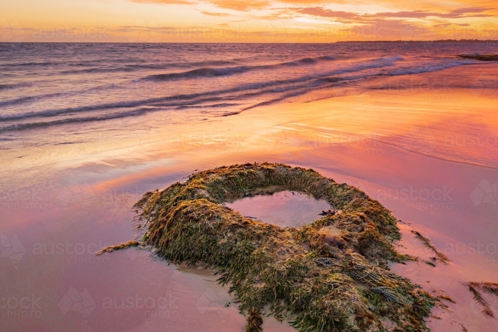 A dramatic sunset reflecting on a wet sandy beach around a solitary circular rock pool - Australian Stock Image