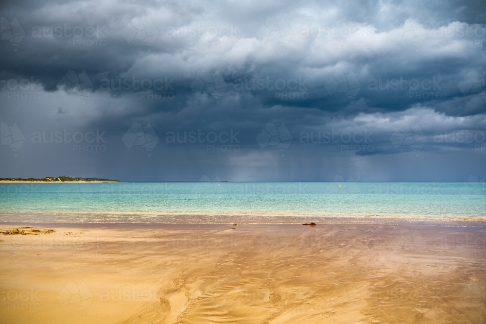 A small stream flowing out to sea over a sandy beach with dark storm clouds and rain over the ocean - Australian Stock Image