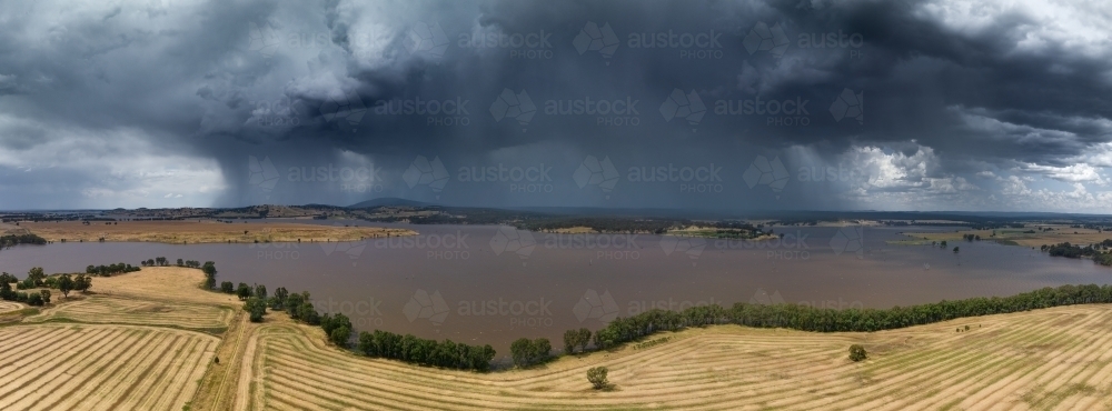 Aerial panoramic view of a rain falling from a large storm cloud over a reservoir alongside farmland - Australian Stock Image