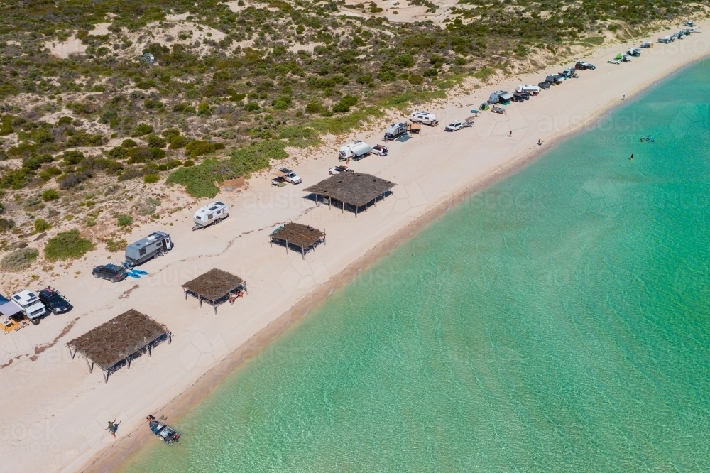 Aerial view of caravans camped along a white sandy beach with beach shelters and calm blue water - Australian Stock Image