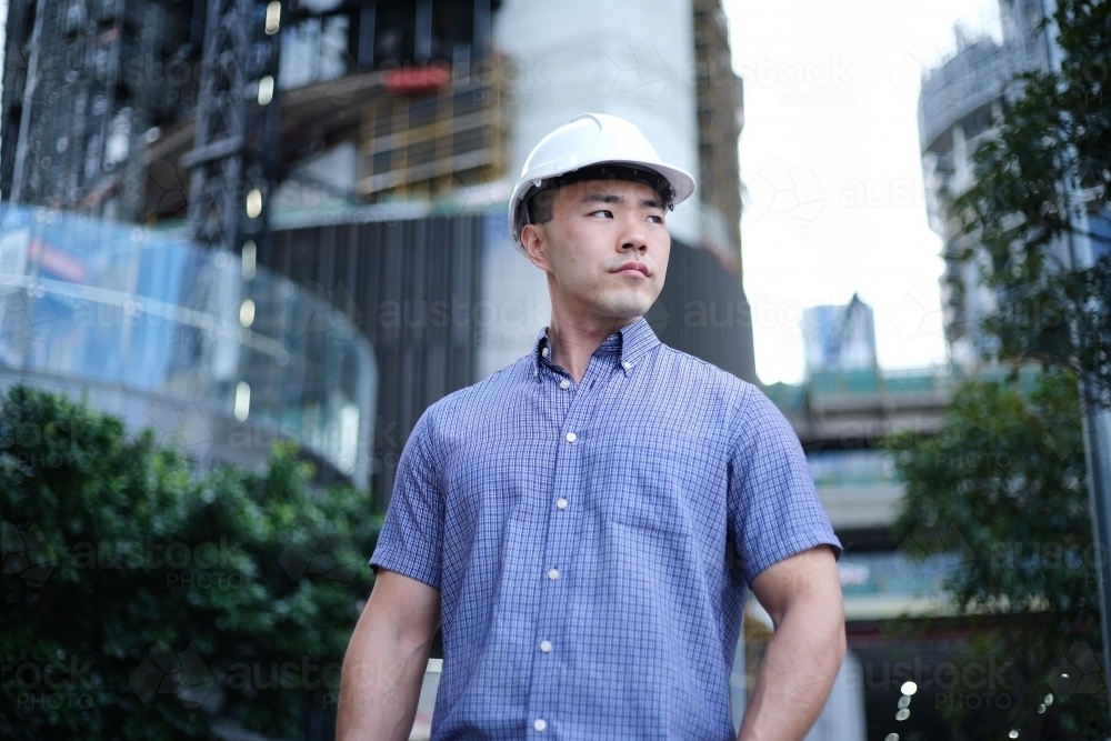 Asian man wearing hard hat with city background - Australian Stock Image
