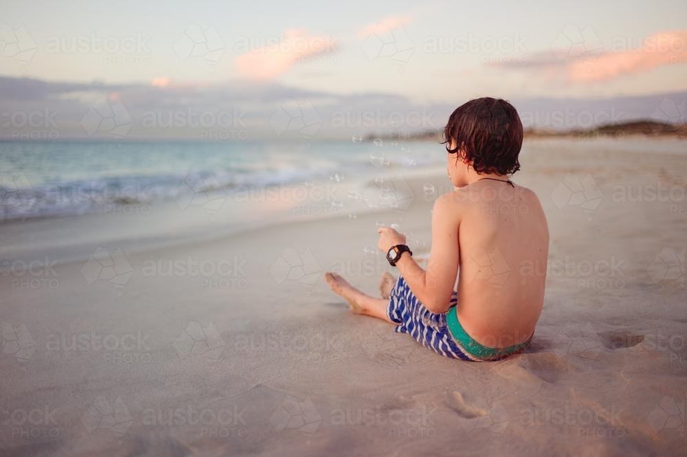 Boy sitting on a beach blowing bubbles on a warm summer evening - Australian Stock Image