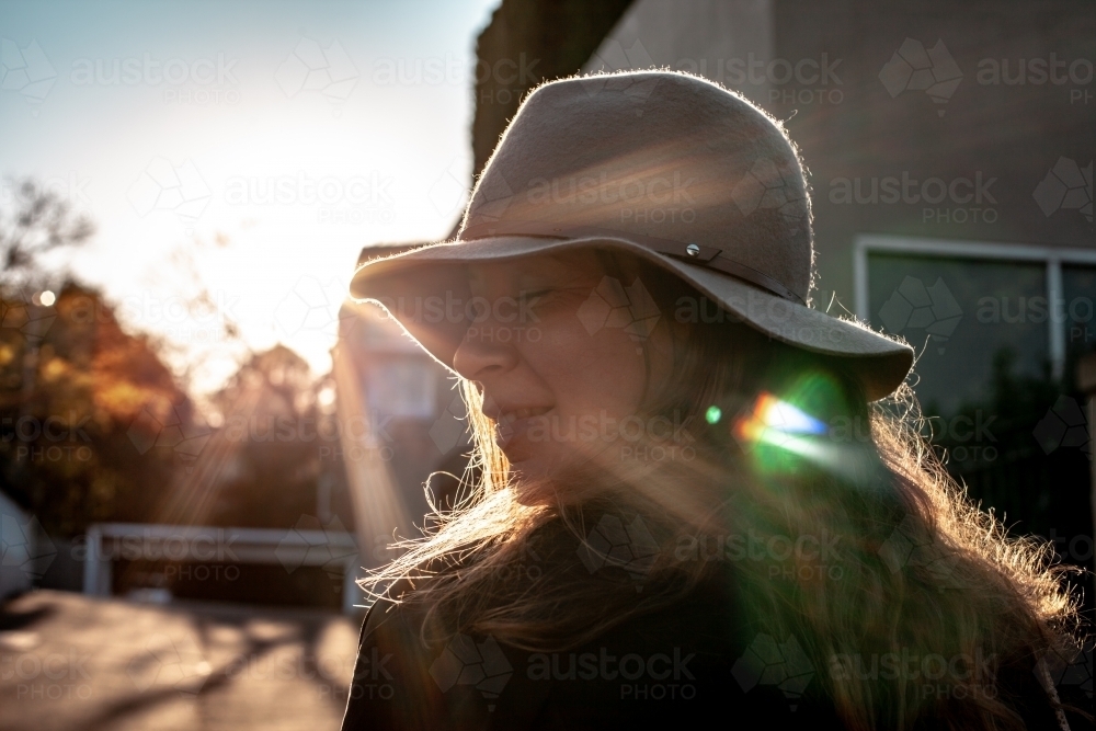 close up shot of woman with eyes closed wearing a hat and a house and sunlight in the background - Australian Stock Image