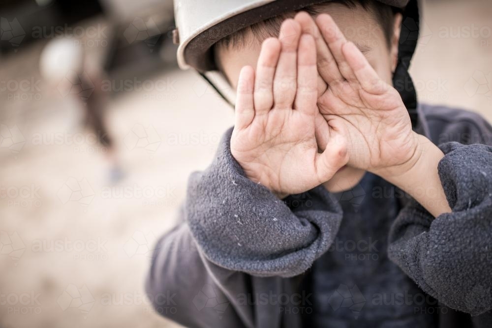Cute mixed race boy hides behind his hands outside wearing a riding helmet - Australian Stock Image