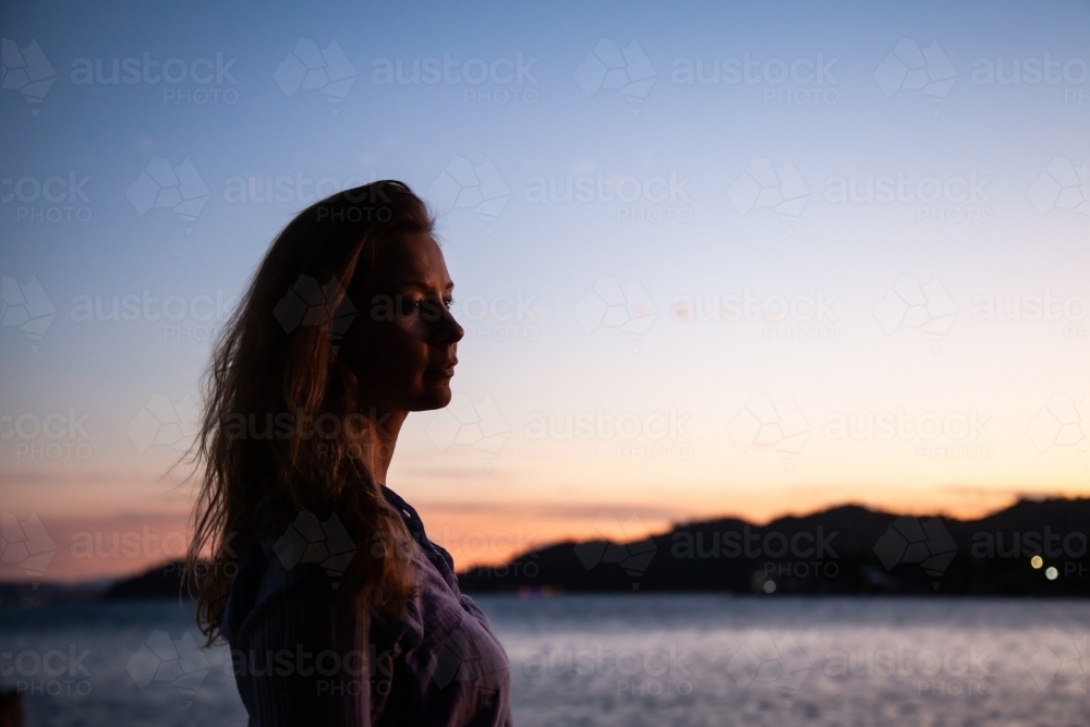 horizontal shot of a silhouette of a woman by the beach with mountains and sunset in the background - Australian Stock Image
