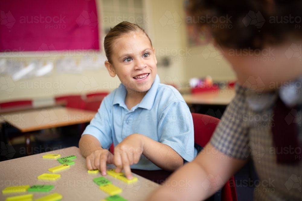 Indigenous girl primary school student sitting smiling at her friend working with word tiles - Australian Stock Image