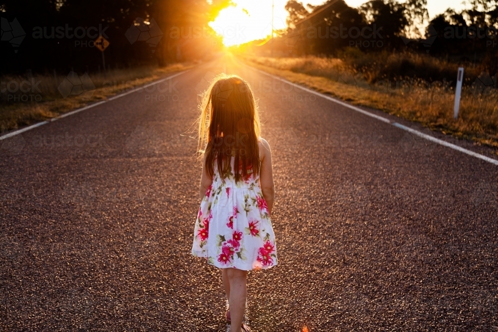 Little girl walking down road towards sunset in glowing golden light - Australian Stock Image