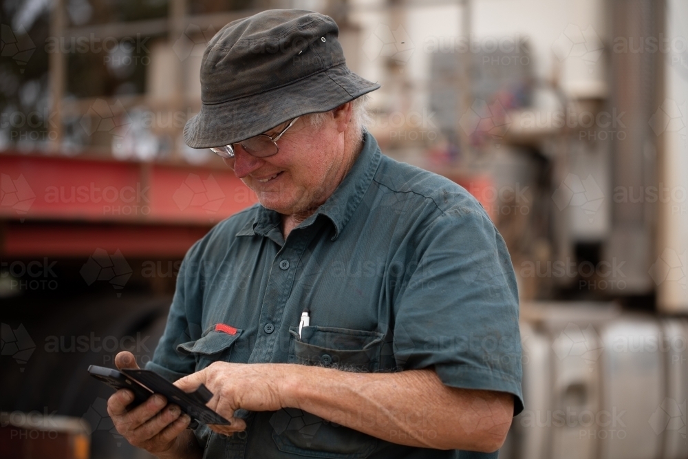 older worker wearing bucket hat using smartphone near machinery - Australian Stock Image