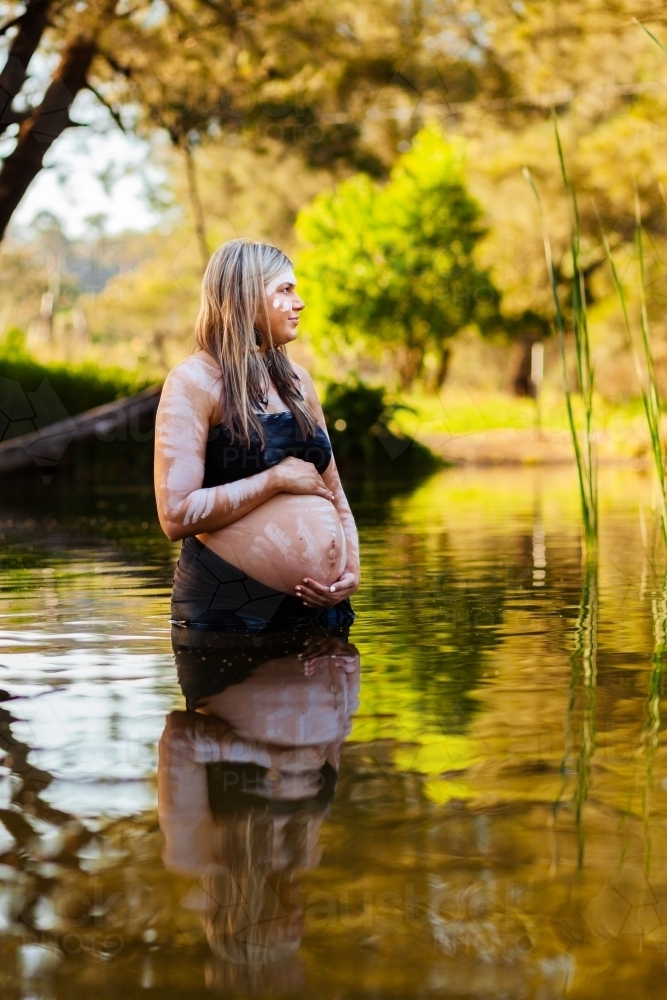 Pregnant Aboriginal Australian woman in traditional ochre body paint in rippling water - Australian Stock Image