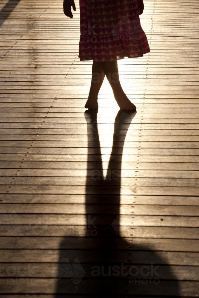 silhouette of a young girl standing on a sunlit deck with crossed legs - Australian Stock Image