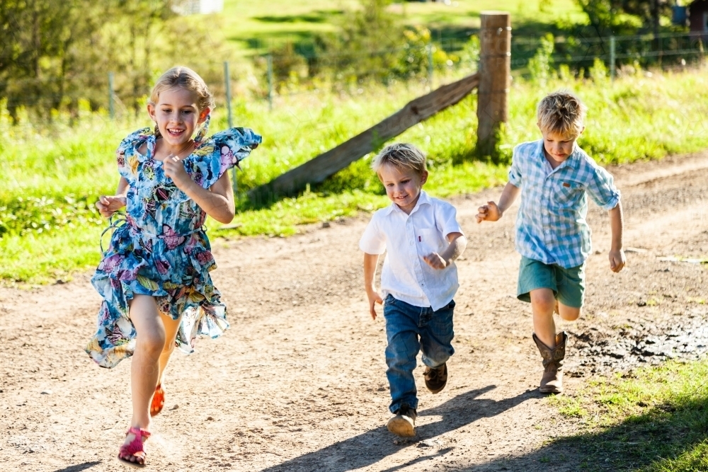Three siblings running down the farm driveway, playing a game together - Australian Stock Image