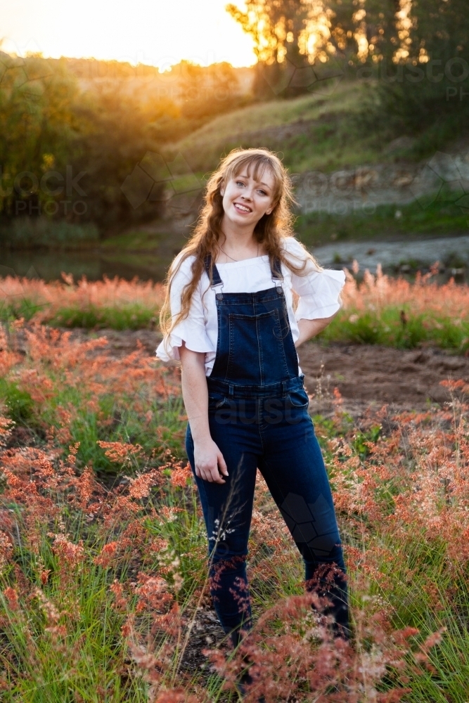 Young adult standing outside by river at sunset in overalls - Australian Stock Image