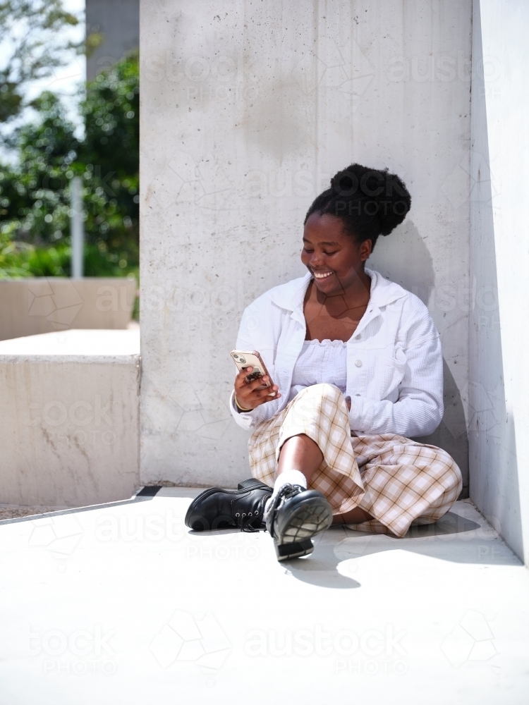 Young African woman using a phone sitting against a concrete wall - Australian Stock Image