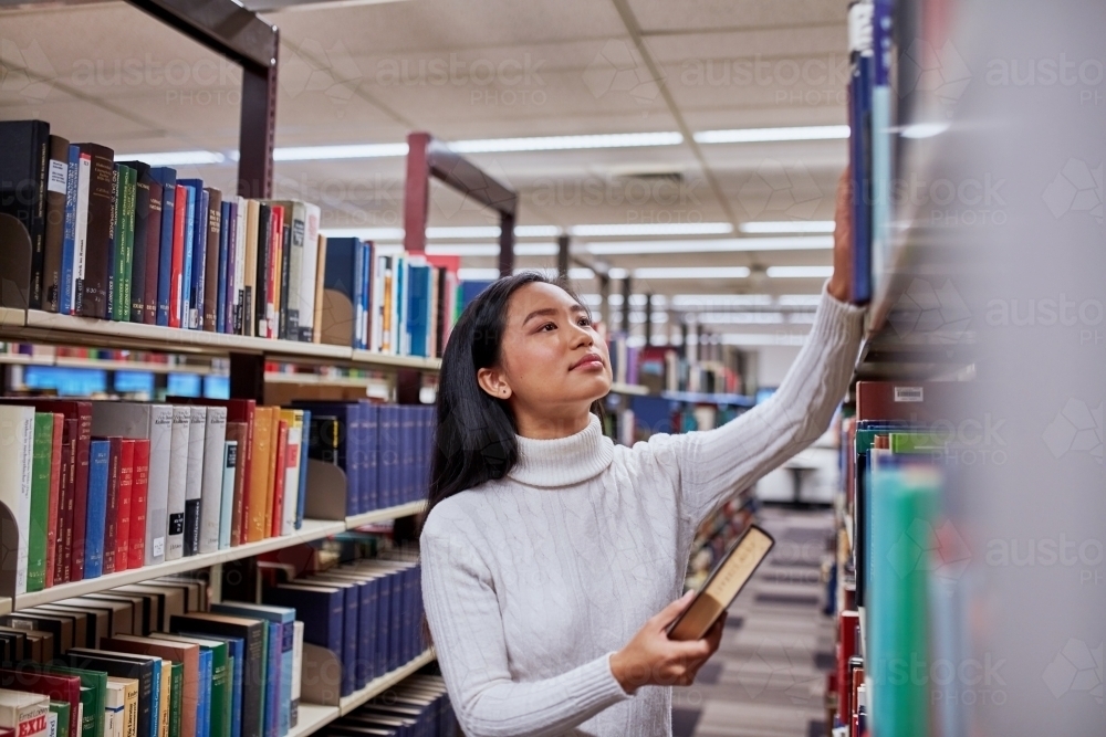 Young Asian student standing in aisle at university library - Australian Stock Image