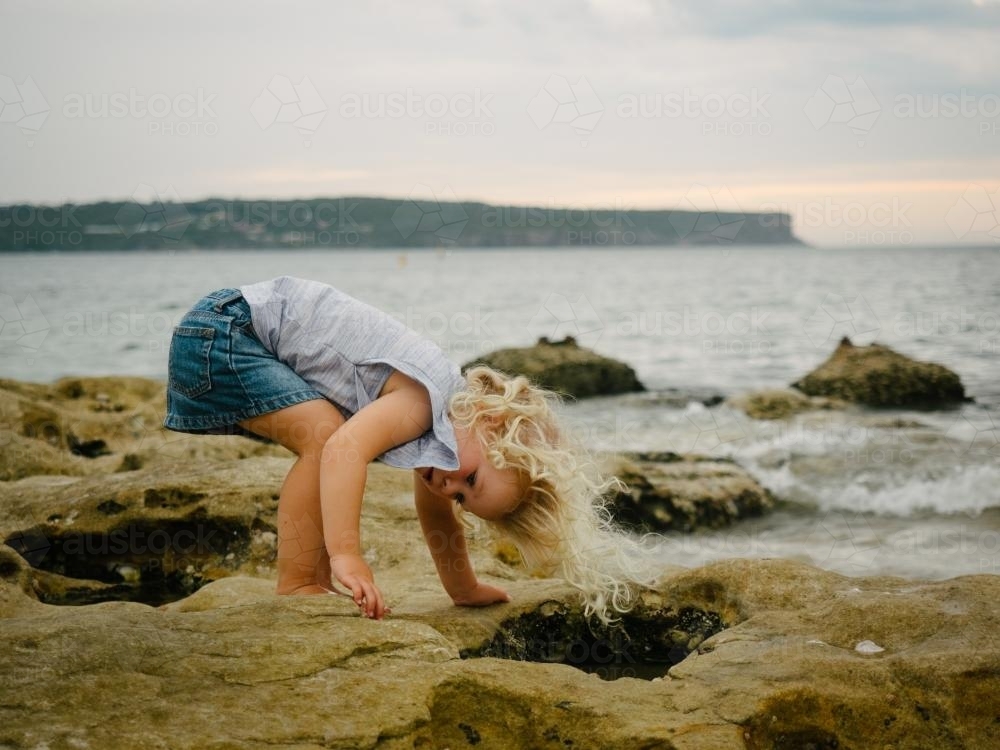 Young girl bending to look in rockpools at the beach - Australian Stock Image