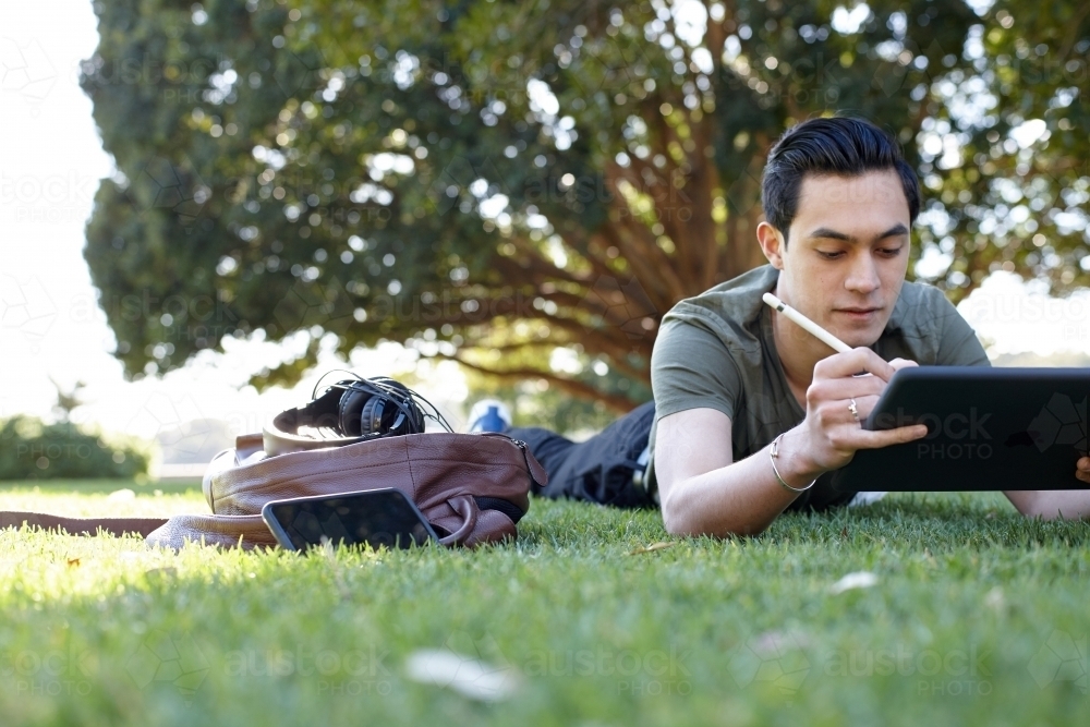 Young man with dark hair writing on his device at park - Australian Stock Image
