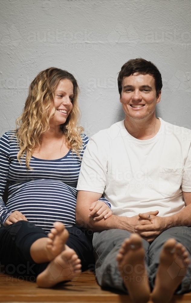 Young pregnant couple sitting against white wall - Australian Stock Image