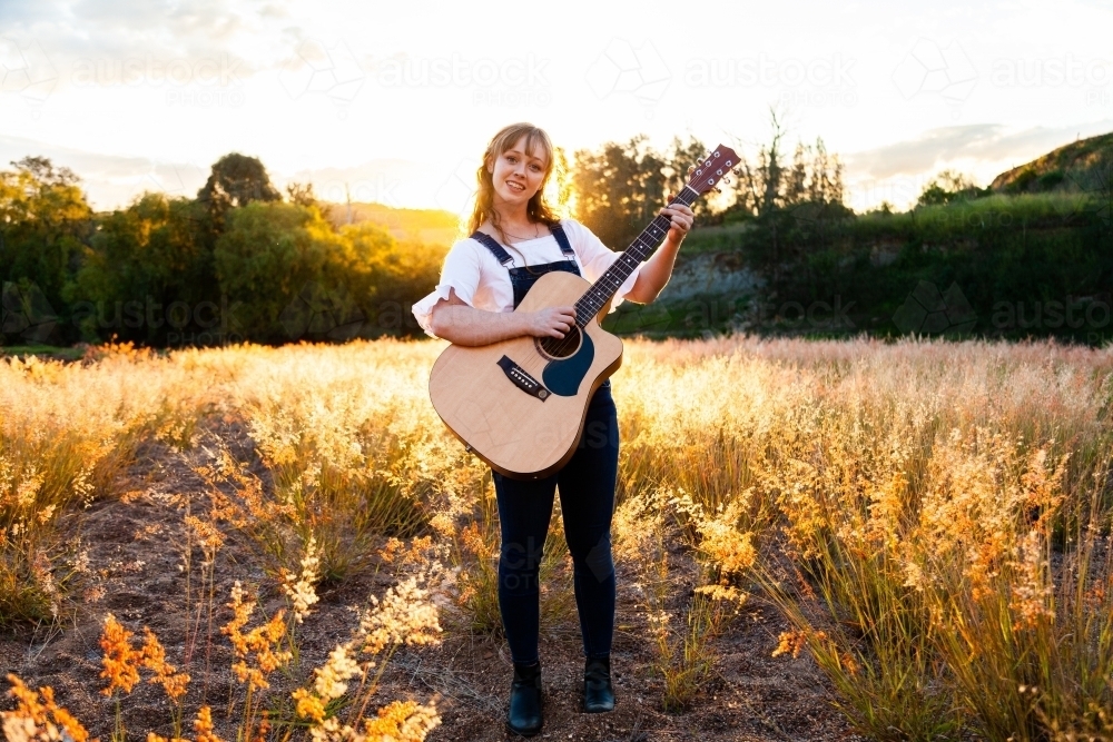 Young woman playing guitar in grass backlit by golden light - Australian Stock Image