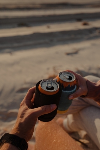 A couple cheersing two cans of drink at sunset on the beach.