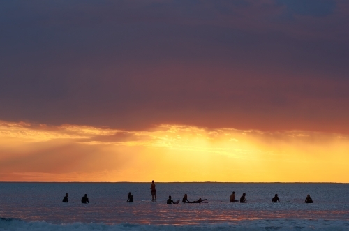 A group of surfers waiting for the next wave, as the sun sets in the background