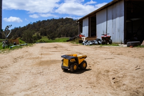 A small, metal, yellow and black toy dump truck, sits central on a gravel road on a farm.