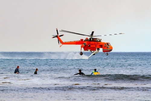 A water-bombing helicopter filling its water tanks from the ocean as surfers watch off Coledale, NSW