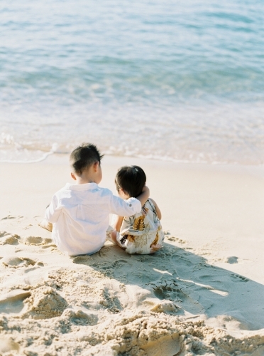 A young boy with his arm around his baby sister sitting on the sand at the beach