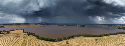 Aerial panoramic view of a rain falling from a large storm cloud over a reservoir alongside farmland