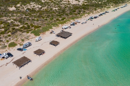 Aerial view of caravans camped along a white sandy beach with beach shelters and calm blue water