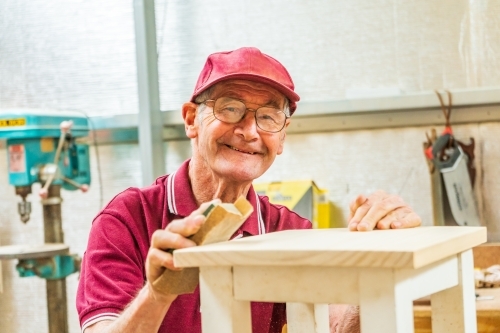 An elderly handyman smiling over the top of a piece of woodwork.