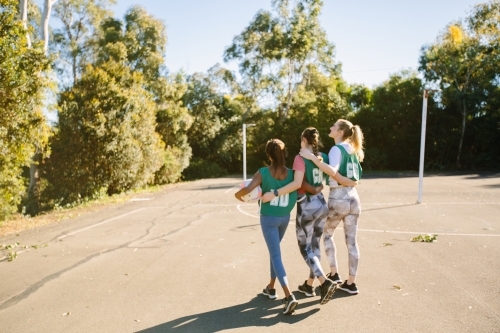 back shot of three young female athletes walking in a field on a sunny day with trees and clear sky
