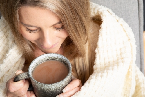 Blonde young lady wrapped in cream blanket hugging a mug