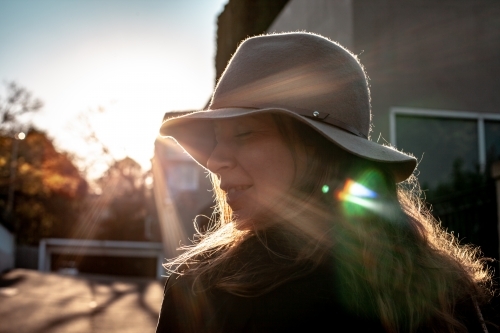 close up shot of woman with eyes closed wearing a hat and a house and sunlight in the background
