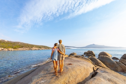 couple with arms around each other standing on granite rock looking out to sea