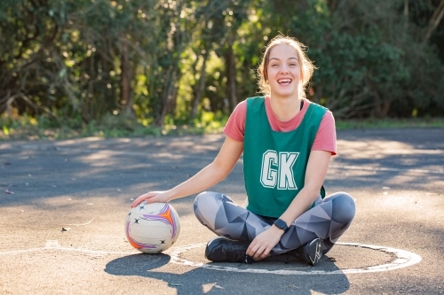horizontal shot of a smiling young woman sitting on the ground with one hand on top of a net ball