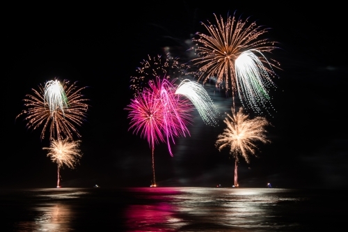 Multi coloured fireworks exploding over an ocean in the night sky.