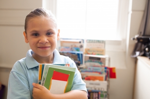 Proud indigenous girl primary school student hugging a pile of books smiling in a classroom