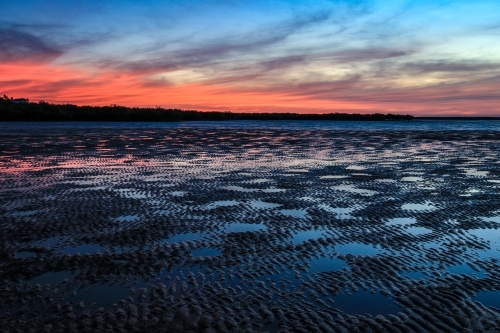 Reflection of sky in pools of water in mud flats beneath colourful sky