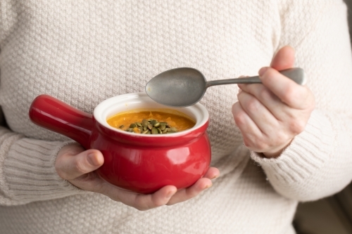 small red bowl of pumpkin soups in lady's hands with spoon