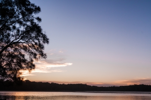 The sun sets behind the silhouette of a tree on the left overlooking a large, smooth lake.