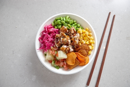top shot of a bowl of chicken vegetable salad with wooden chopsticks on top of a grey background