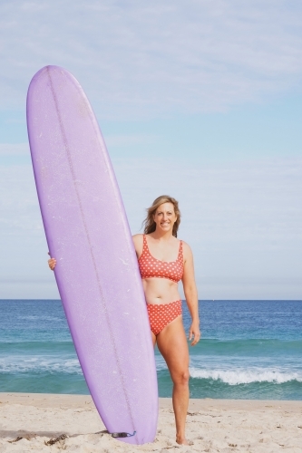 Woman standing on a beach wearing polka dot bikini holding longboard surfboard