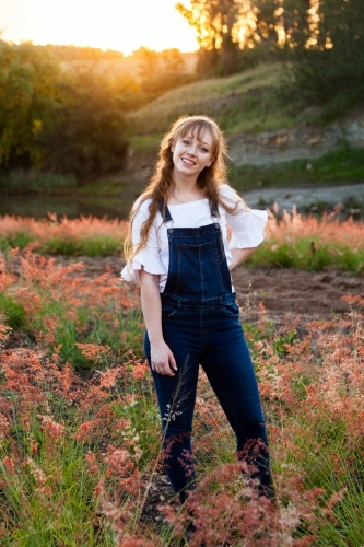 Young adult standing outside by river at sunset in overalls