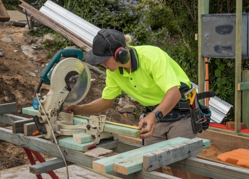 Young apprentice builder cutting timber with drop saw on residential construction site