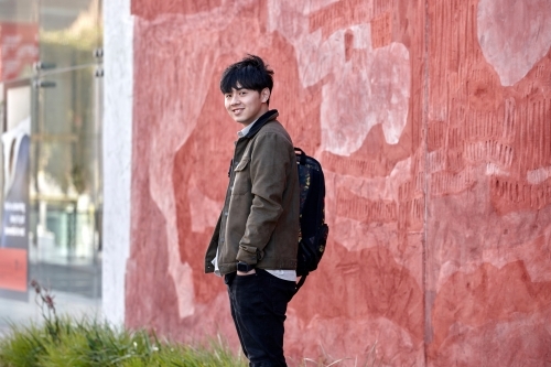 Young Asian university student smiling standing next to red wall on-campus