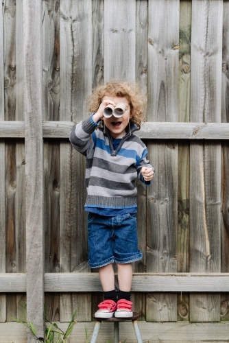 Young boy standing on a stool playing with cardboard toilet roll binoculars in the backyard