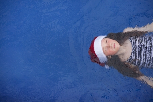 Young girl floats in a pool wearing a festive christmas santa hat