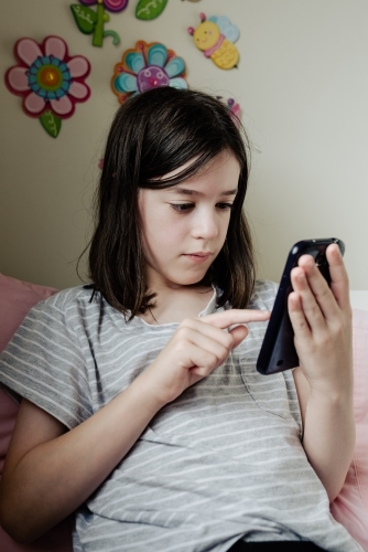 Young preteen girl in her bedroom sitting on bed texting chat messages on mobile phone / smartphone