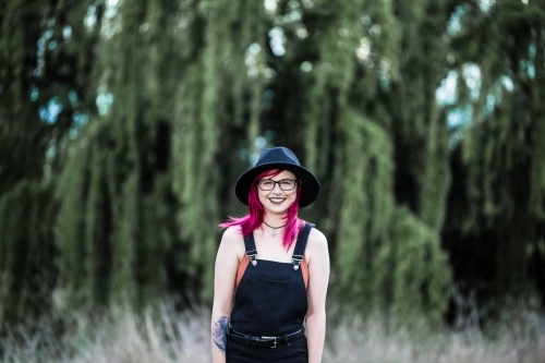 Young woman with pink hair smiling in front of willow tree