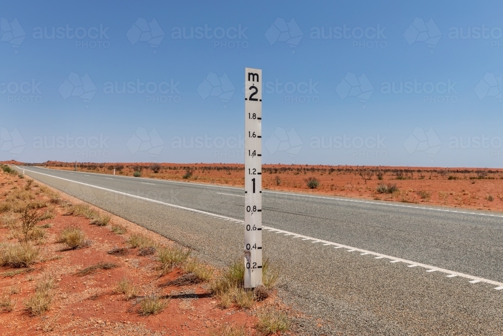 2 metre flood level marker on the edge of a long road crossing red, dry flood plain - Australian Stock Image